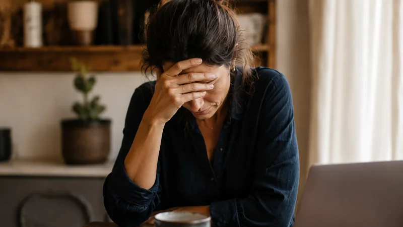 Femme assise à une table, la main posée sur le front, devant un ordinateur et des documents